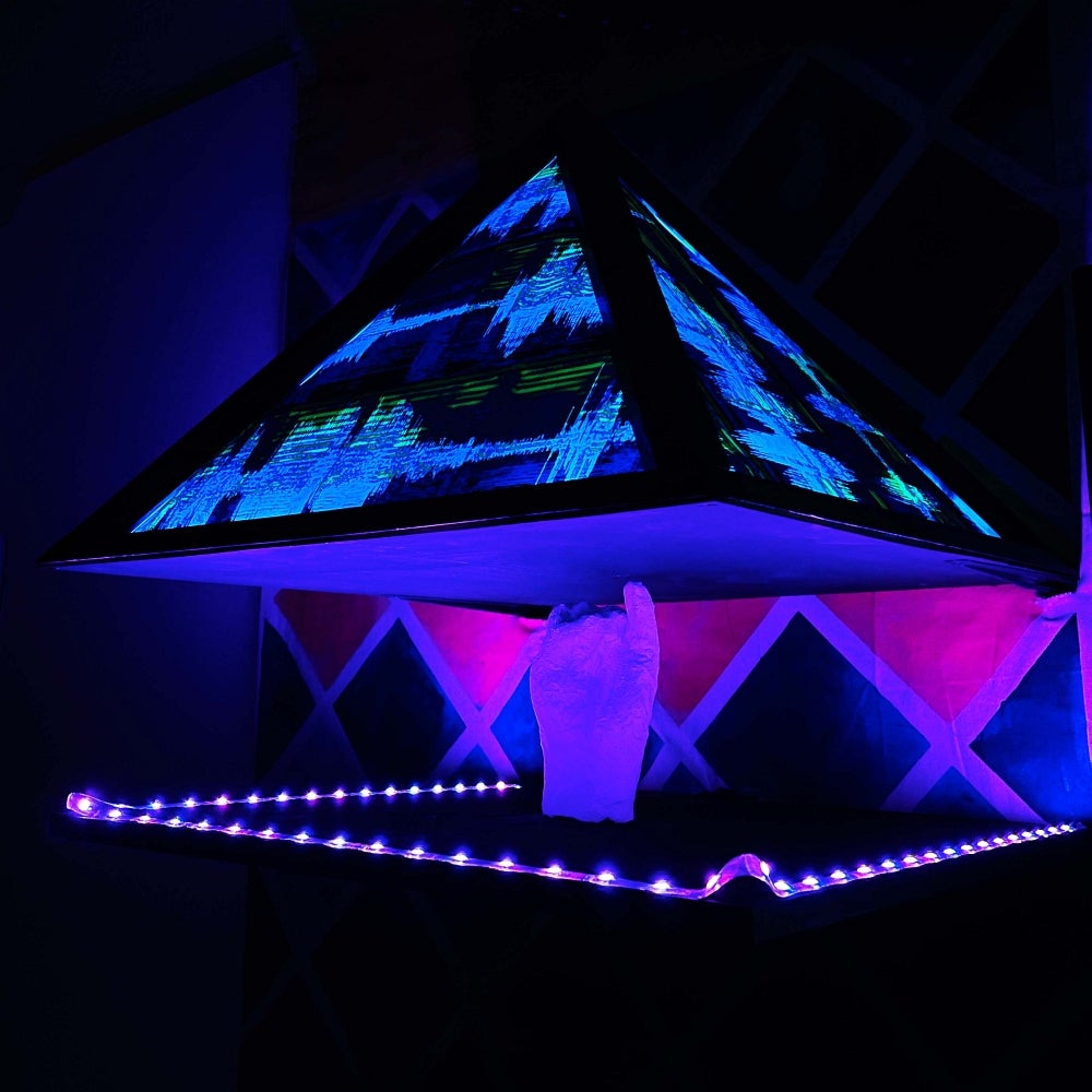 Artwork in glowing shades of blue and magenta, a pyramid shape with digital interference patterns is held up by a hand.