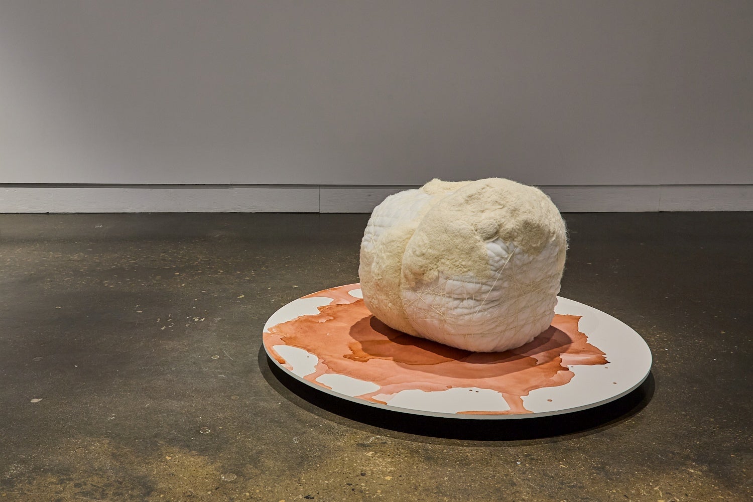 Large white and cream, spherical, fabric sculpture on a circular platform decorated with an oxidized stain.