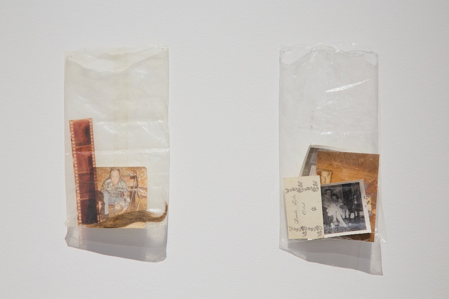 Two clear plastic bags containing old photos, ephemera and a lock of hair, hang on a wall.
