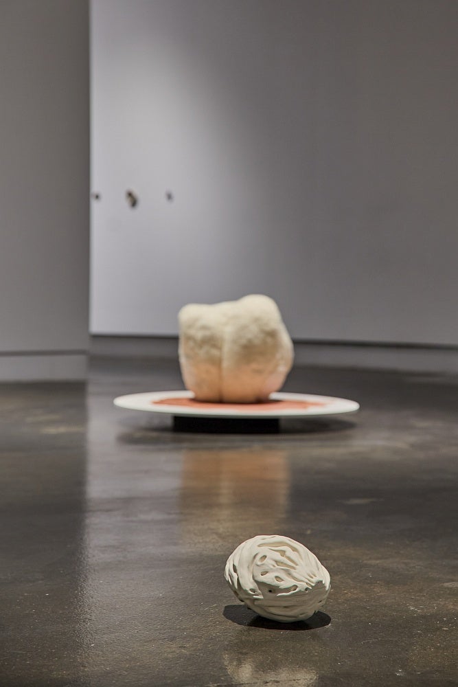 A large off-white porcelain peach pit rests on the floor in front of a spherical fabric sculpture on circular platform.