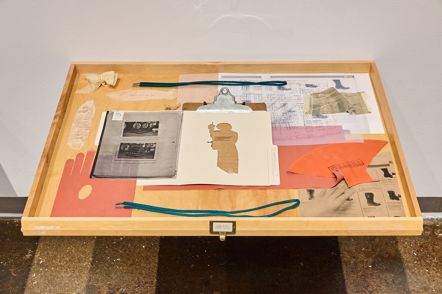 Wooden drawer displaying vintage plans, product catalogue pages, file folder, clipboard, and rubber condoms decorated with text.