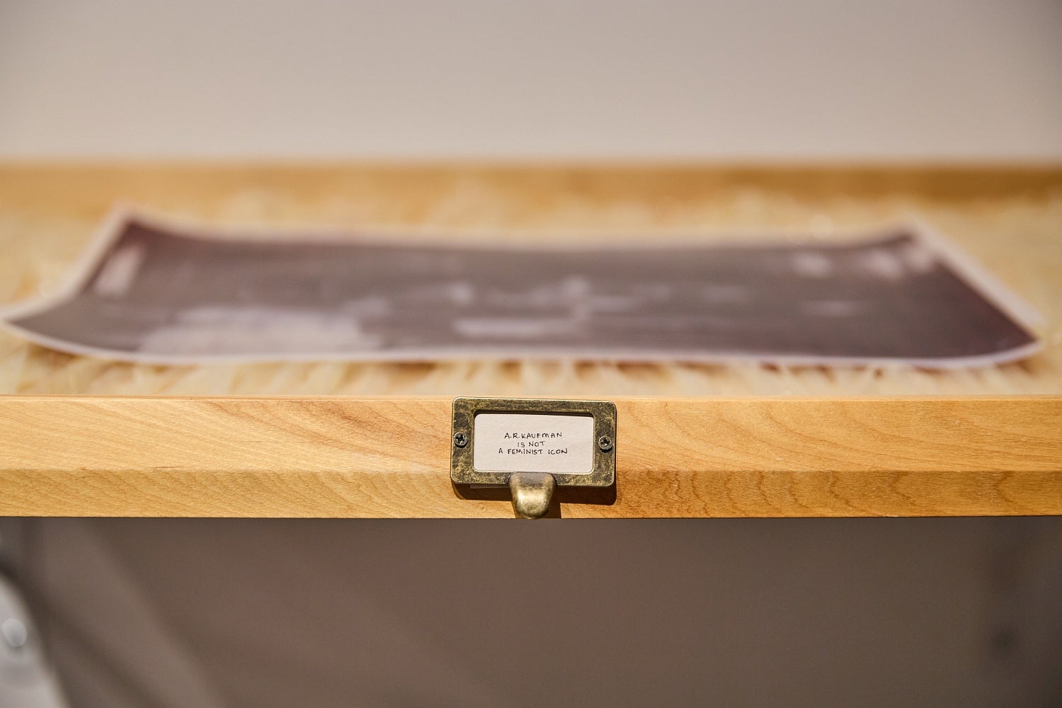Detail of a wooden shelf with a brass drawer pull label reading "A.R. Kaufman is not a feminist icon".