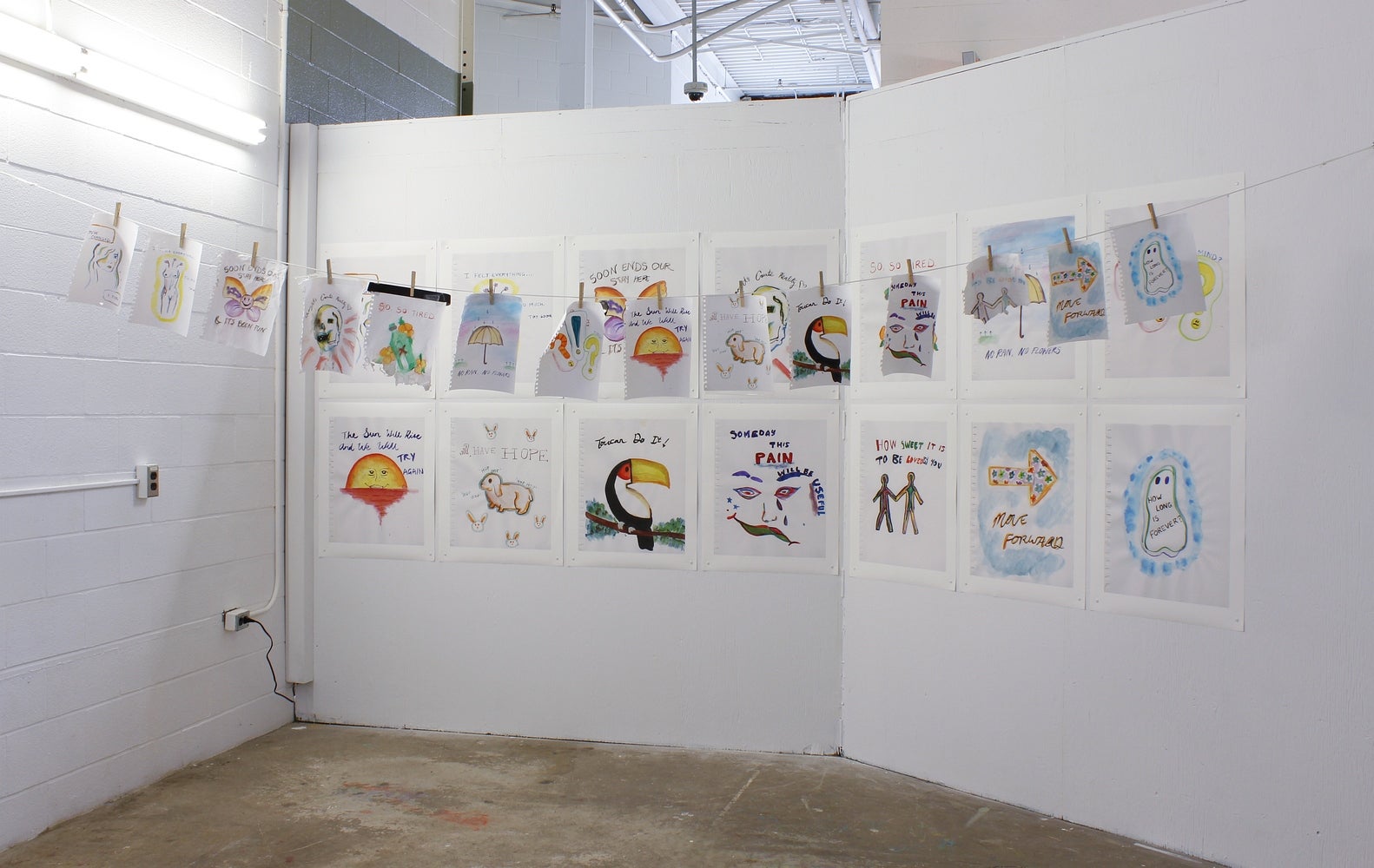 Art installation of watercolours hung in two rows of seven across two walls with a cord hung across the corner and small waterco