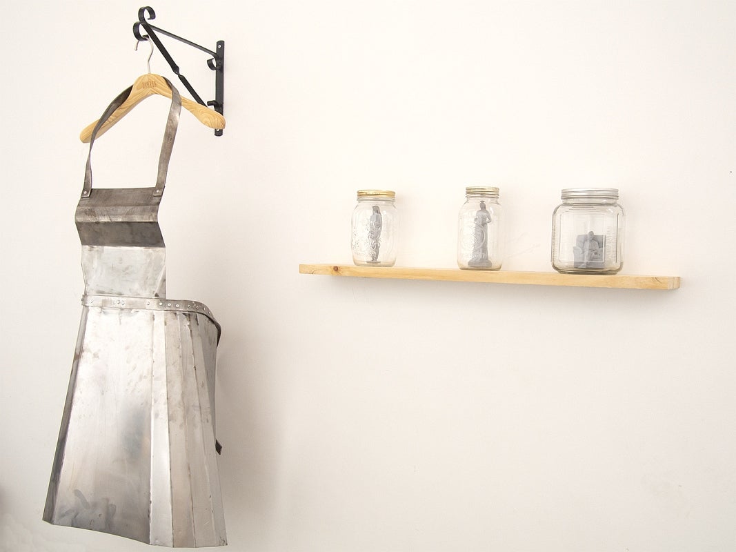 Art installation of an apron made of steel on a coat hanger and a wood shelf with 3 mason jars containing small grey figures.