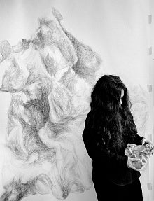 Black and white image of a woman in long hair looking down at a rock-like object in her hands.  Behind her is a large drawing of the same object.