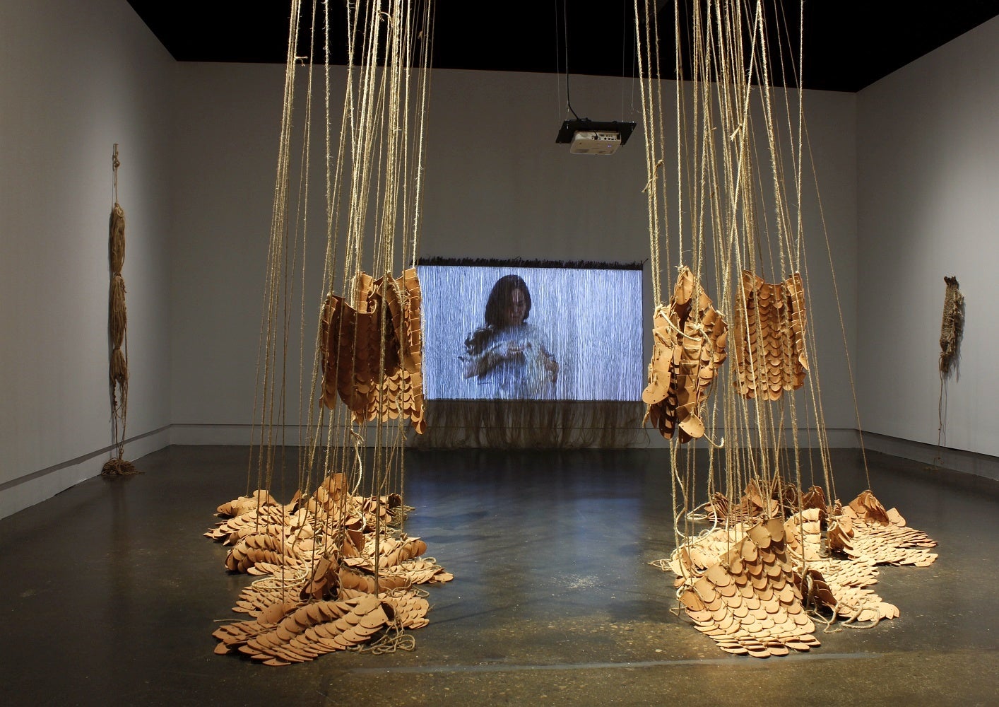 Art gallery with installation of videos and multiple rounded clay tiles bound together using jute hanging from the ceiling and piled on the floor.