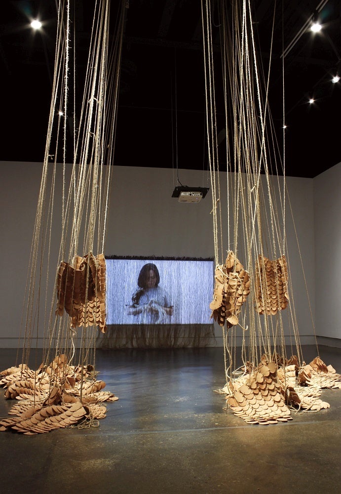 Art gallery with installation of videos and multiple rounded clay tiles bound together using jute hanging from the ceiling and piled on the floor.