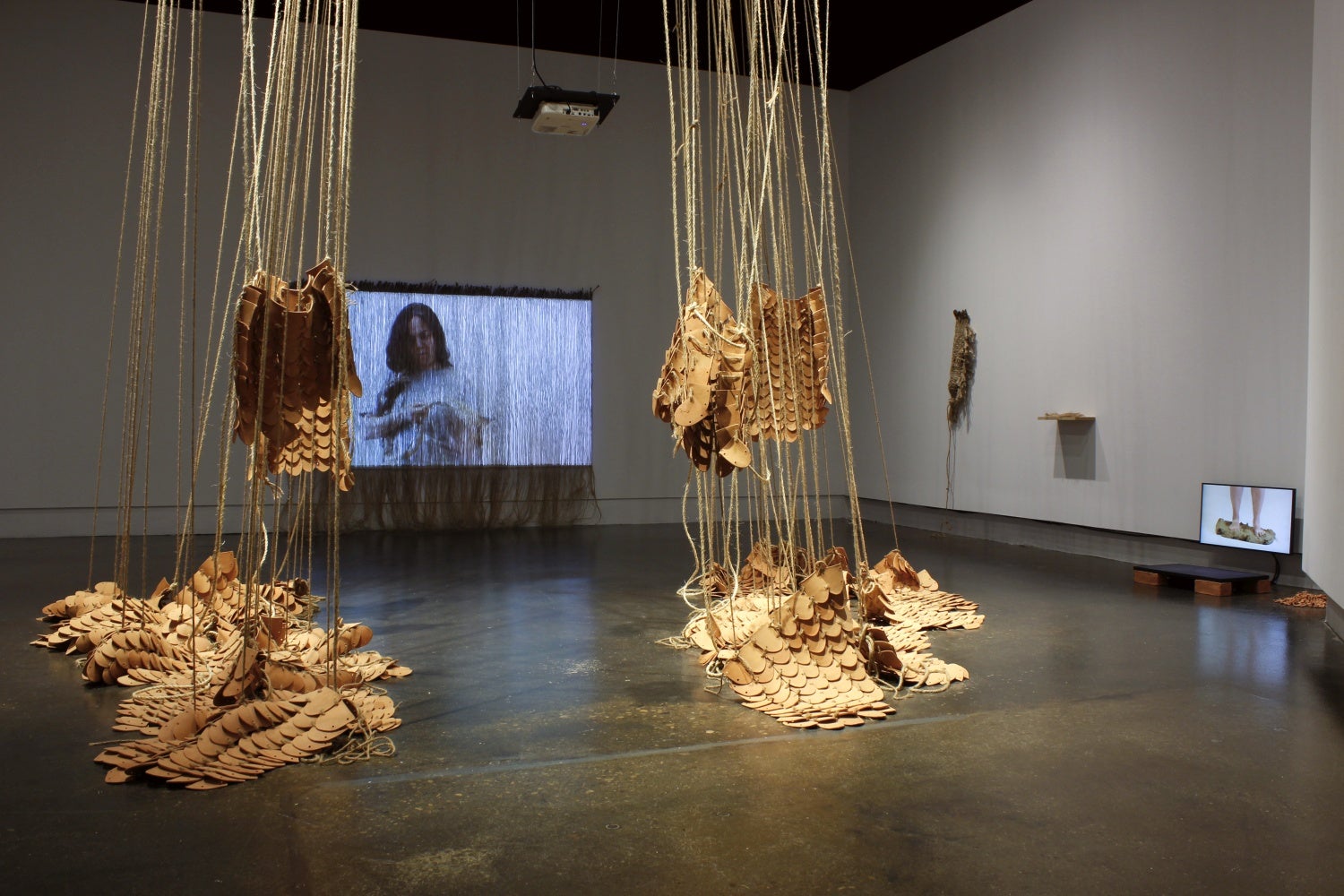 Art gallery with installation of videos and multiple rounded clay tiles bound together using jute hanging from the ceiling and piled on the floor.