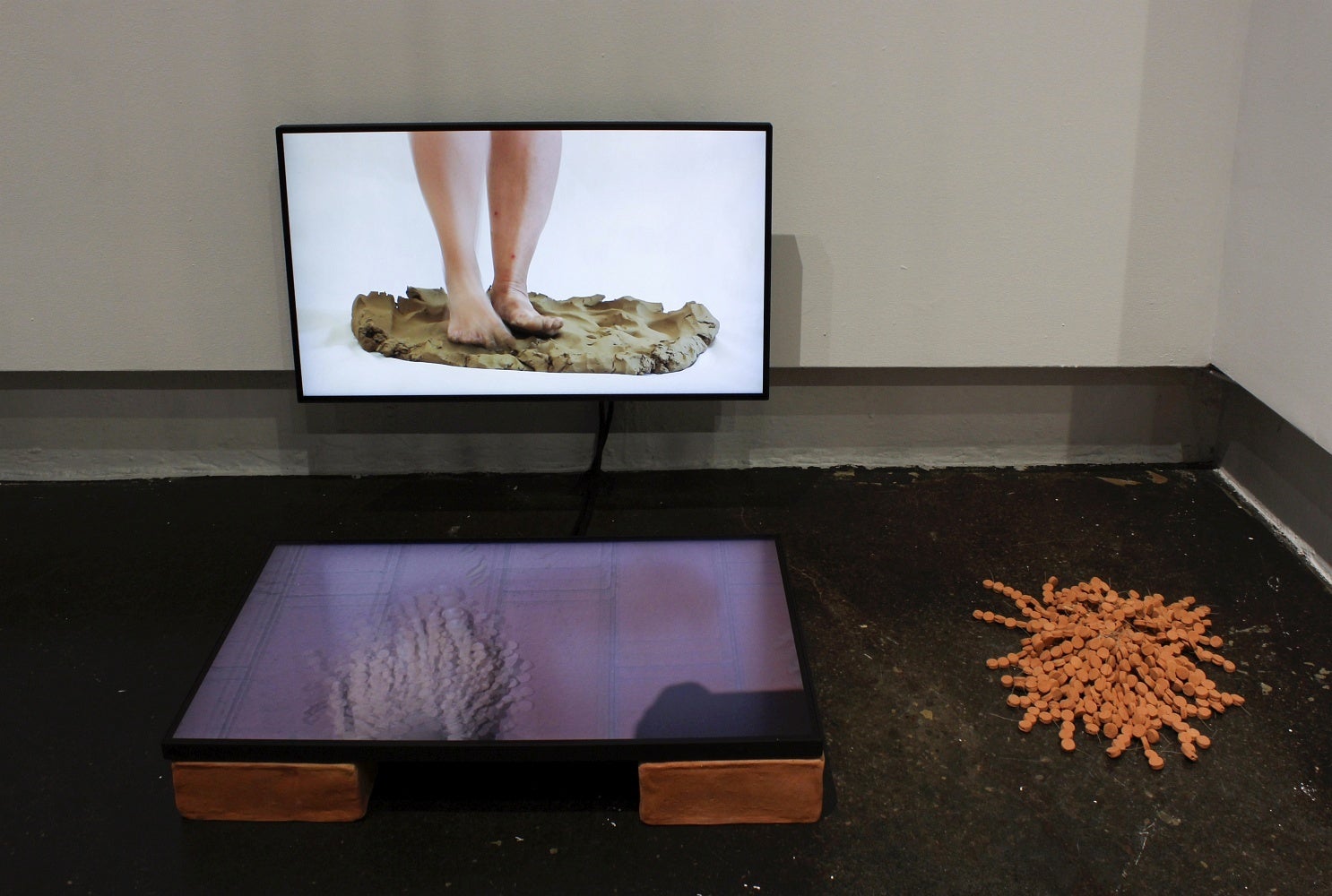Art installation of a videos of feet kneading clay and walking with anklets of clay discs.