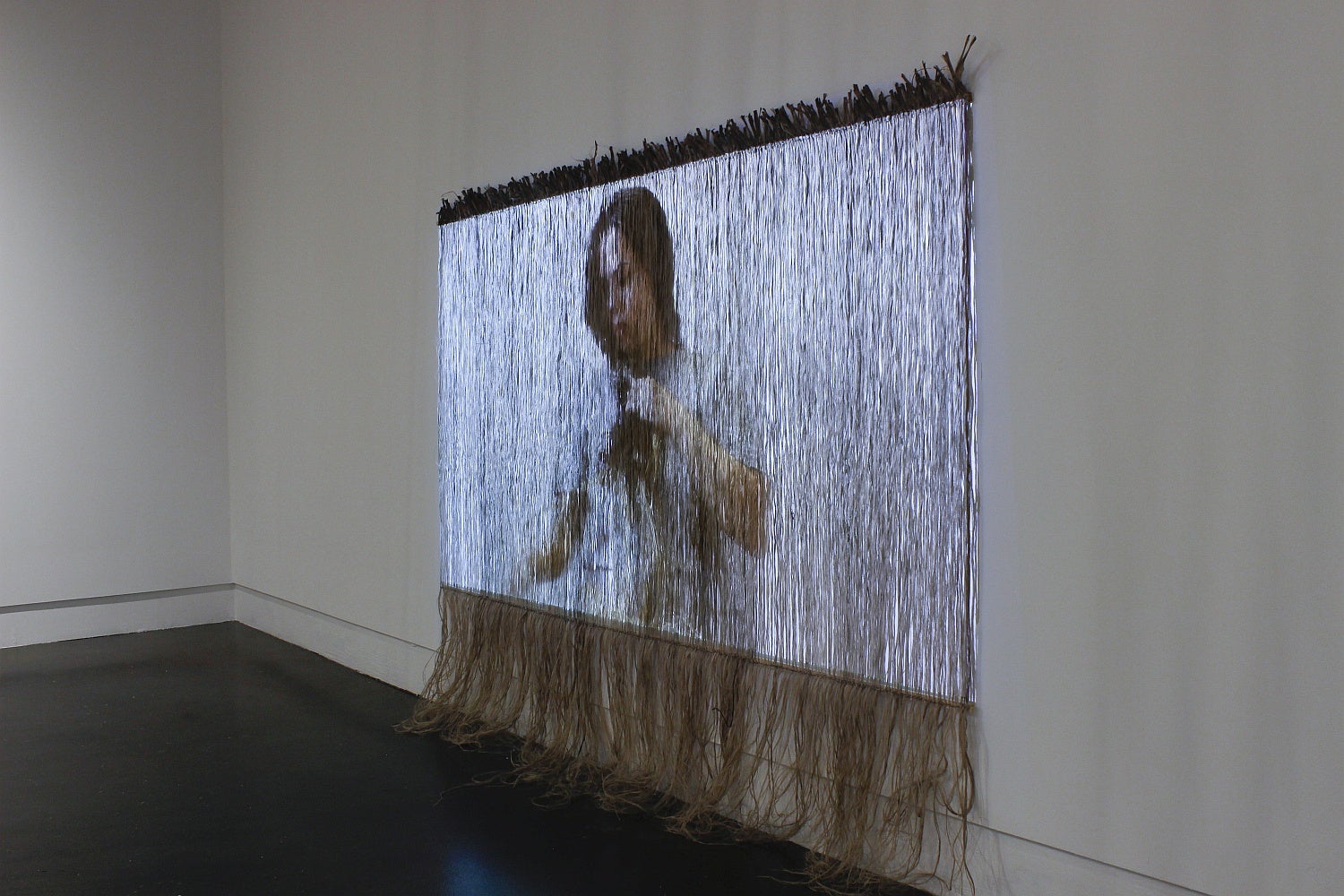 Video of a women braiding rope into her hair, is projected onto a curtain of jute strips.