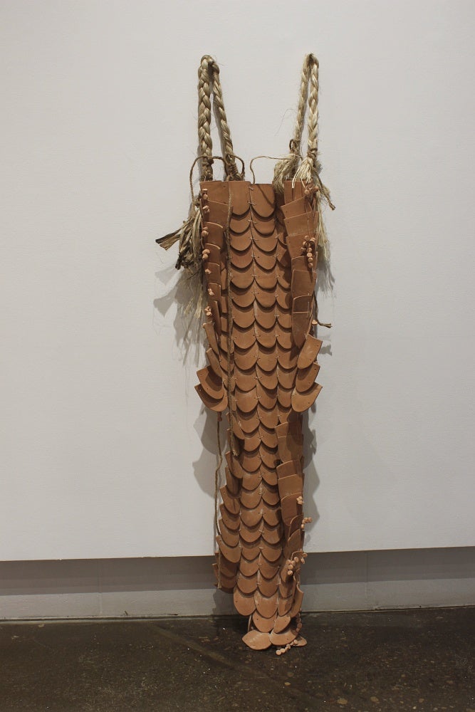 Multiple rounded clay tiles bound together using jute to form a cape, hang on the wall.