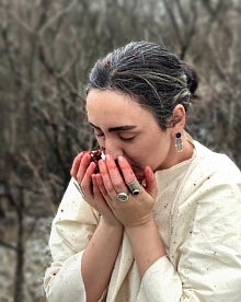 Figure holds their hands, stained red, to their face to smell sumac fruit.
