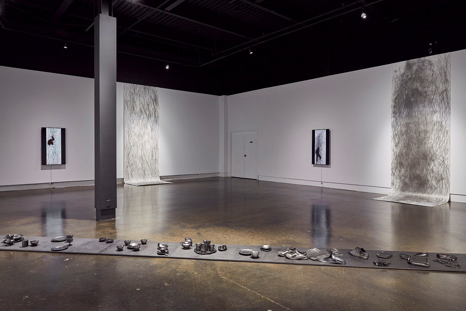 Art gallery exhibition with video monitors, large hanging drawings that drape onto the floor, and a row of blackened objects on the floor.