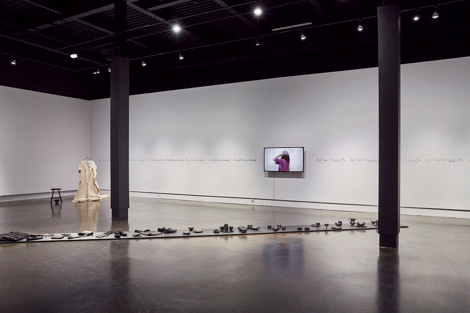 Art gallery exhibition with a video monitor, text running along the wall, a canvas dress hung from the ceiling and a row of blackened objects on the floor.