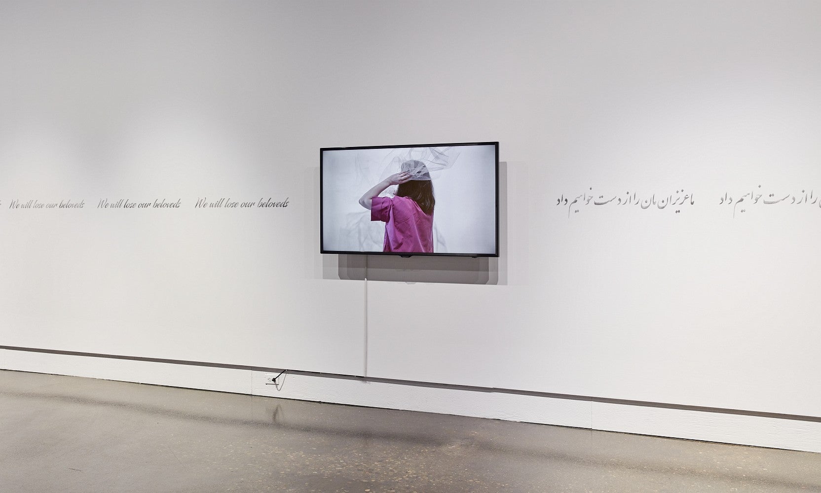 Art gallery exhibition with a video monitor and text in English (We will lose our beloveds) and Arabic running along the wall.