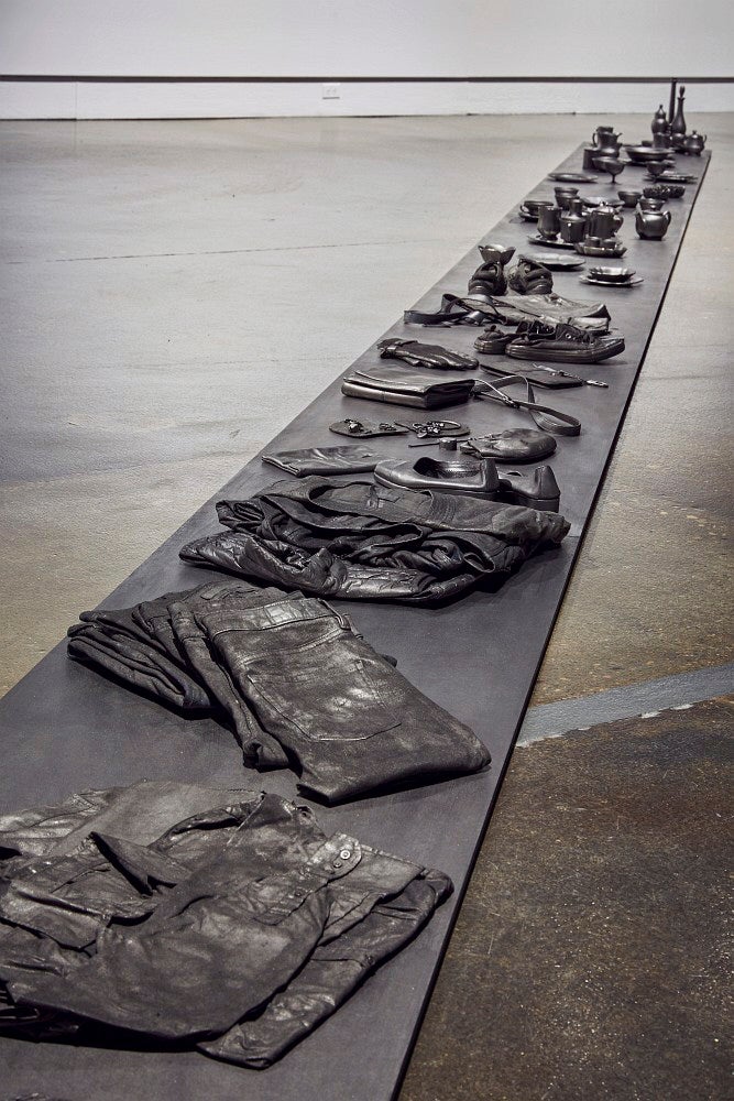 A long line of objects, including folded clothing and dishes, all blackened with graphite, sit on a concrete floor