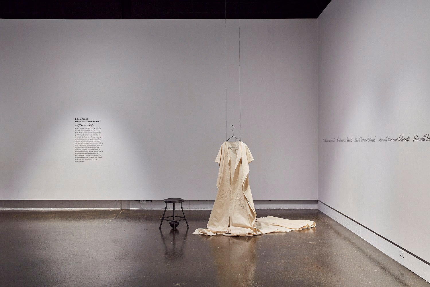 Art gallery exhibition text running along the wall, a canvas dress is hung from the ceiling with a blackened stool beside it.