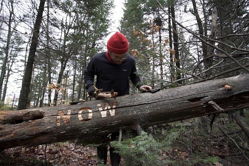 In a wooded area a person in a red toque leans over a fallen tree that has the letter NOW carved in it.