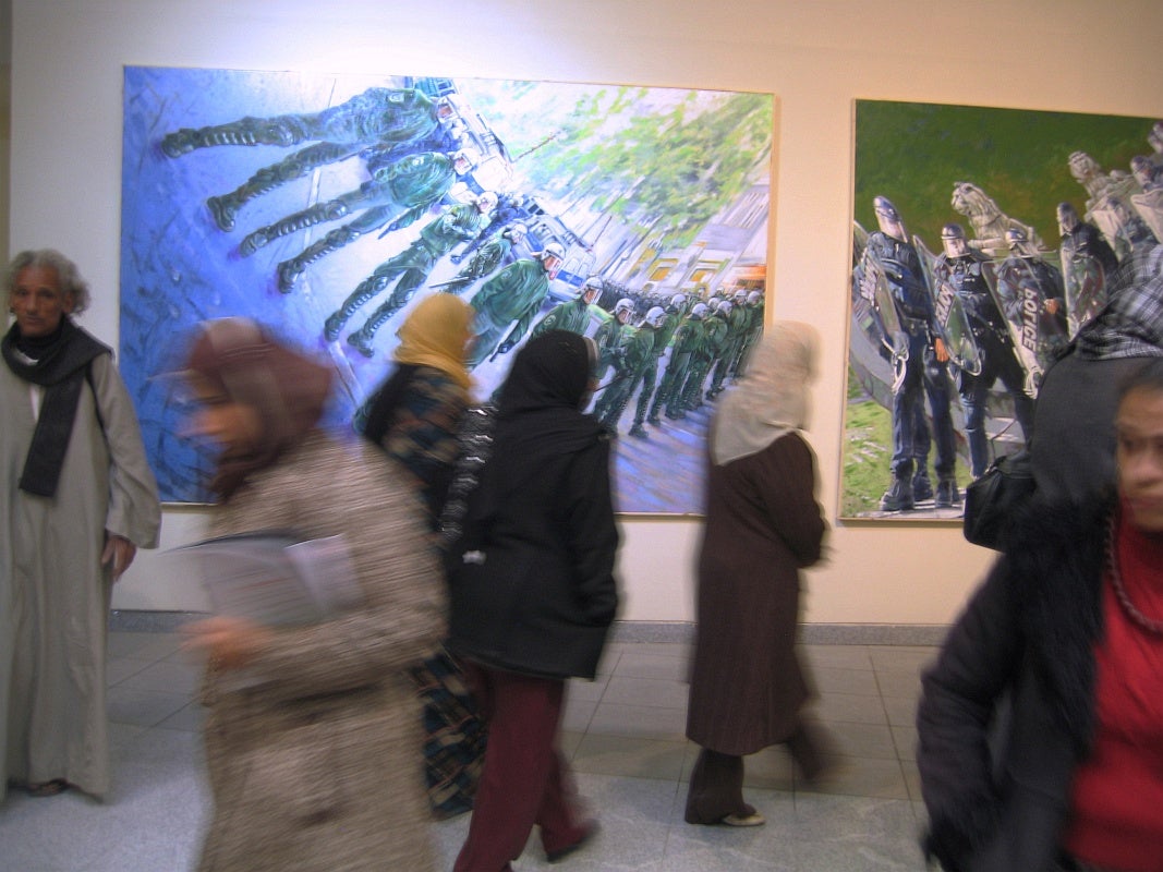 Motion blur showning multiple people, several of them wearing head scarves, moving in front of two paintings each depicting a line of police in riot gear with anamorphic distortion. 