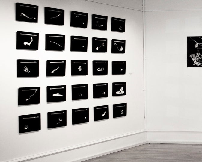 Gallery installation of artworks hung in a 5x5 grid.  Works are black and feature white shapes.