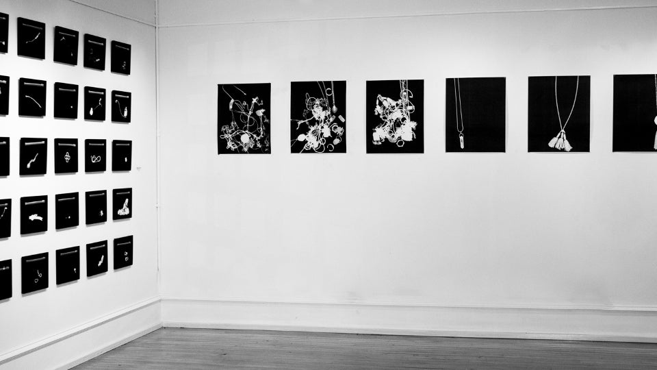 Gallery installation, one wall works in a 5x5 grid, second wall 6 works in a line. Works are black and feature white shapes.