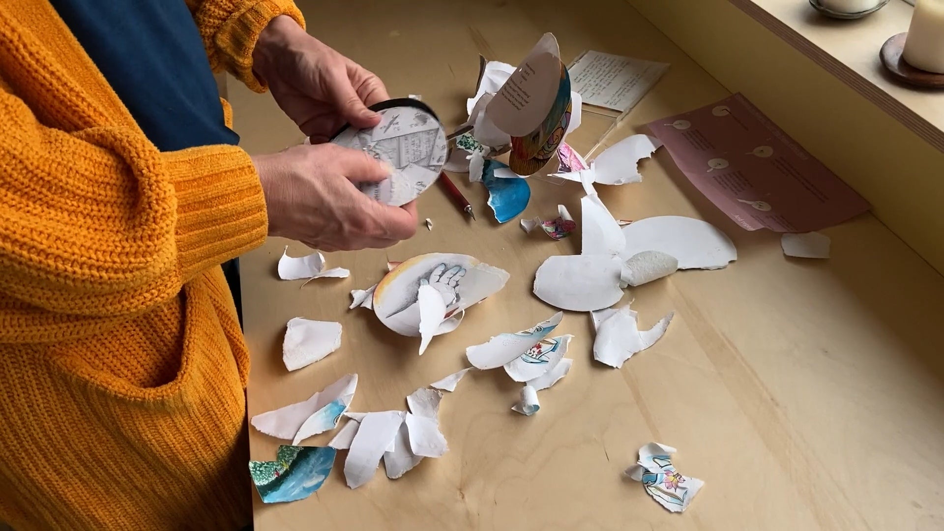 Person holding a circular drawing stands by a table covered with bits of torn drawings.