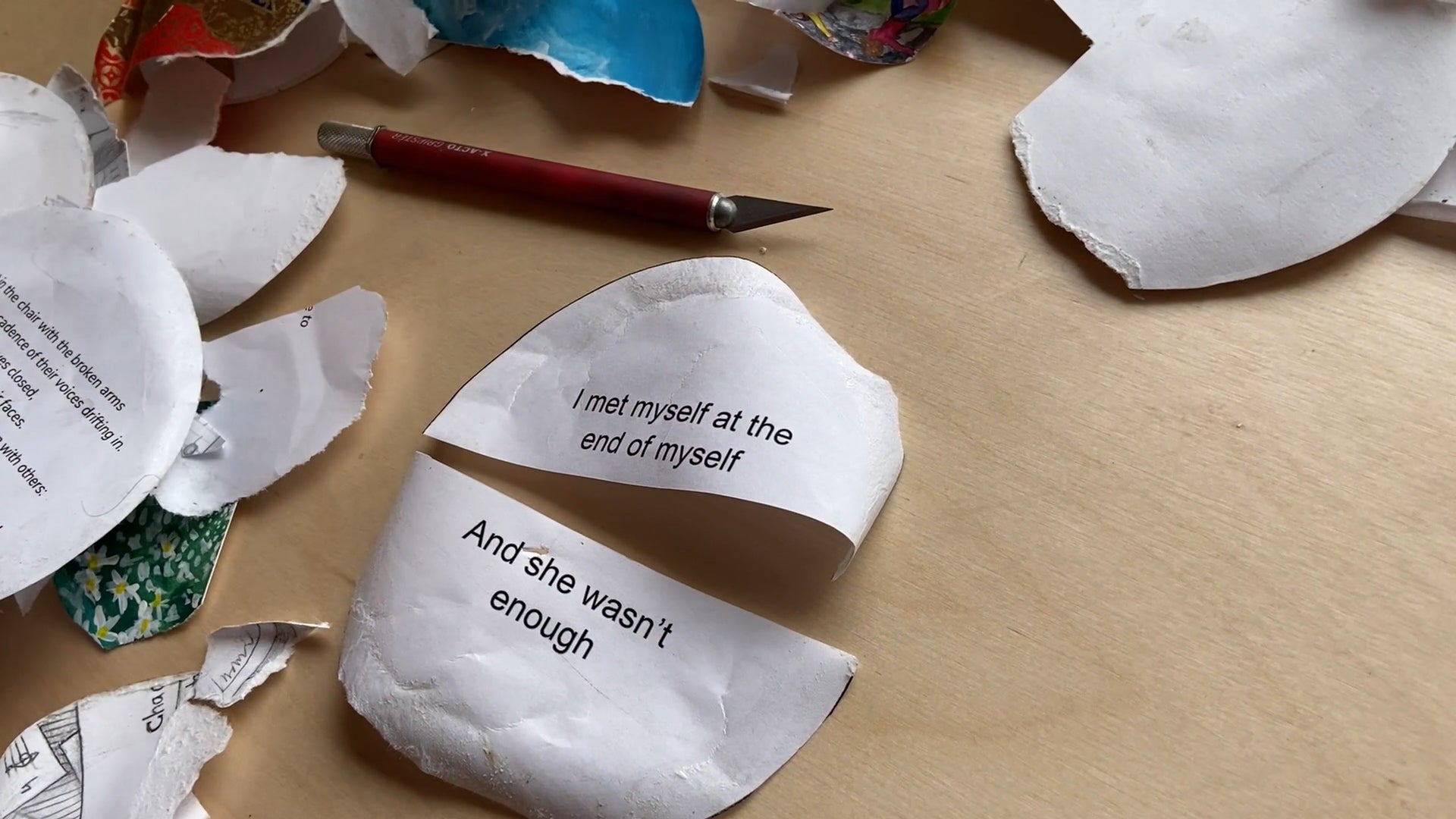 Table covered with bits of torn paper, a knife and a circular paper, cut in half, with text "I met myself at the end of myself a