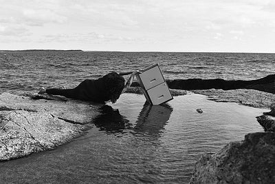 The artist, lying on their side on a rocky tidal lagoon at the ocean's edge, struggles to hang off a precariously balanced two-drawer filing cabinet sitting in the water