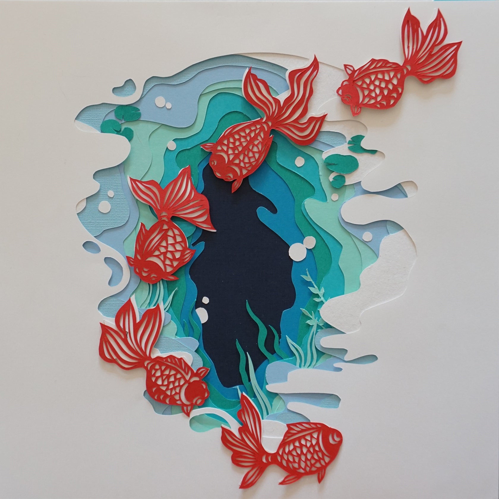 Paper cut-out artwork, white paper cut to reveal pages in shades of blue and green like water, five orange carp swim over page..