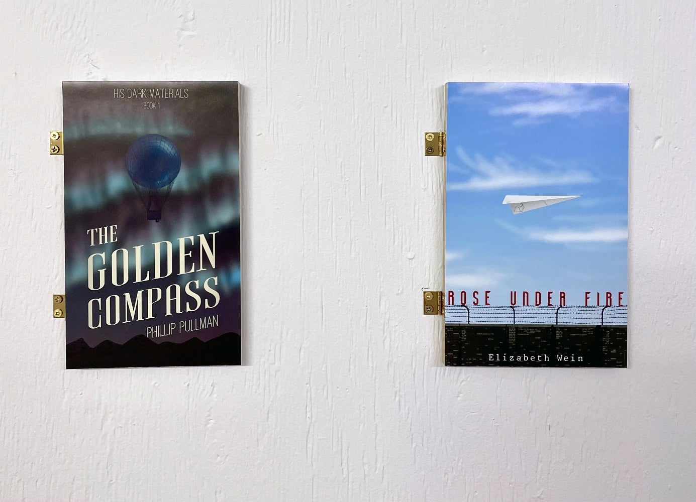 Art installation of 2 book covers attached to the wall with hinges titled "The golden compass" and "Rose under fire"