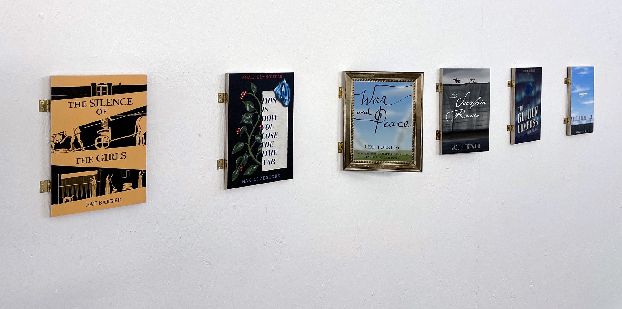 Art installation of 6 book covers attached to the wall with hinges, one is positioned slightly open, away from the wall.