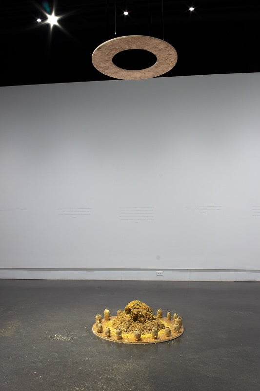 On the concrete floor in the center of the room in a circular wooden base, painted gold and filled with wood-chips surrounded by potatoes. A second chipboard circle is suspended from the ceiling. On the back wall, lines of hand printed text are just barely visible.