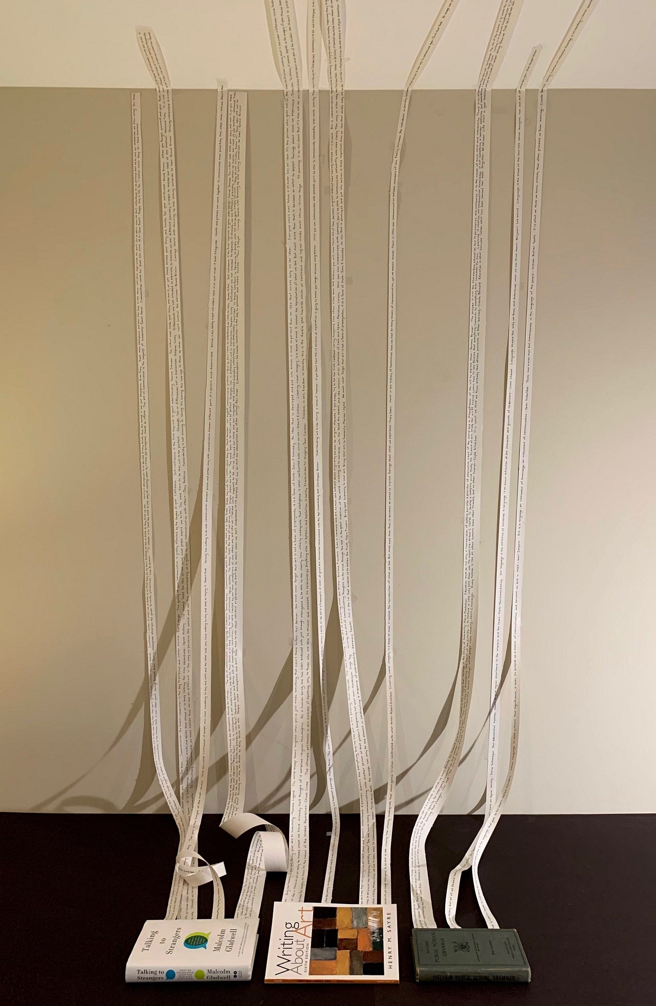 Art installation of three books lying on the floor with strips of paper coming from inside the books and stretched up the wall