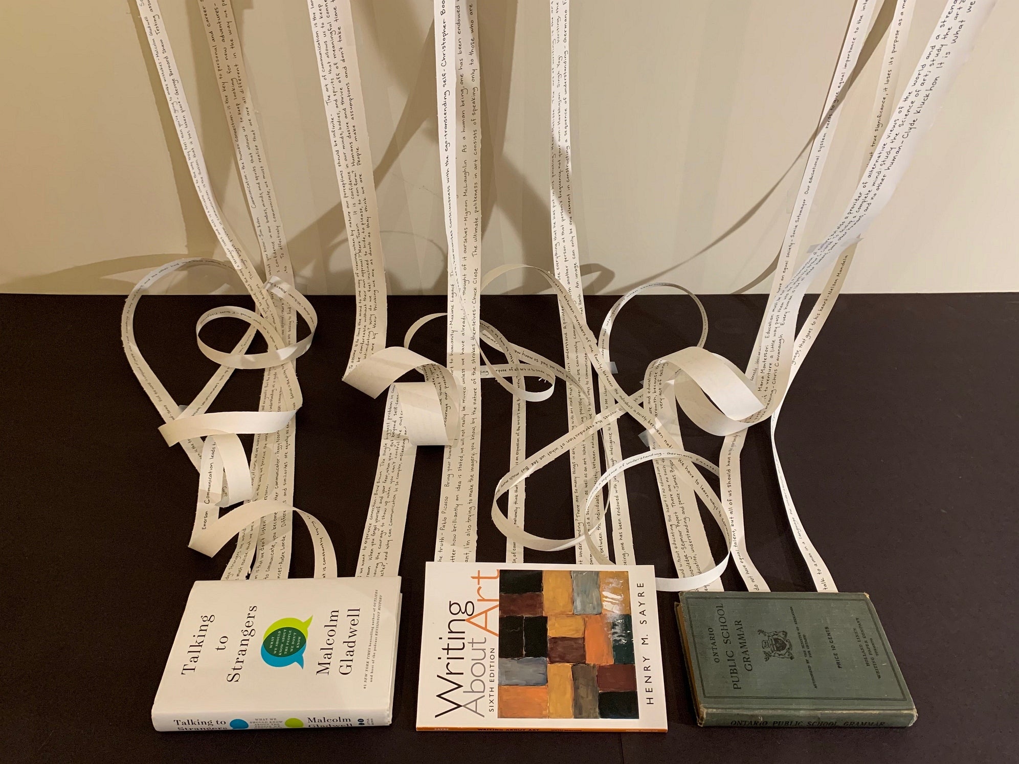Art installation of three books lying on the floor with strips of paper coming from inside the books going up the wall