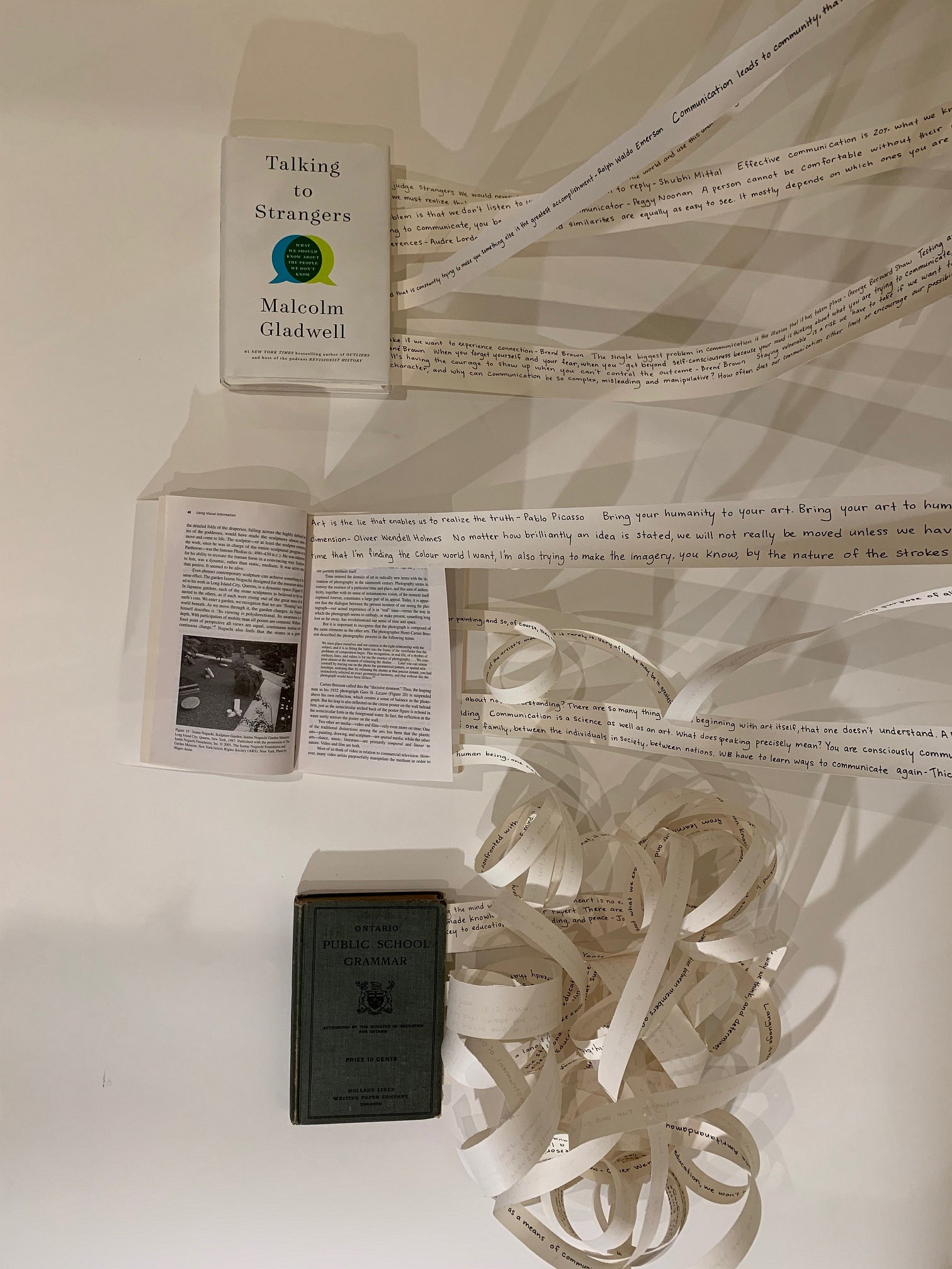 Art installation of three books lying on the floor with strips of paper coming from inside the books and stretched up the wall