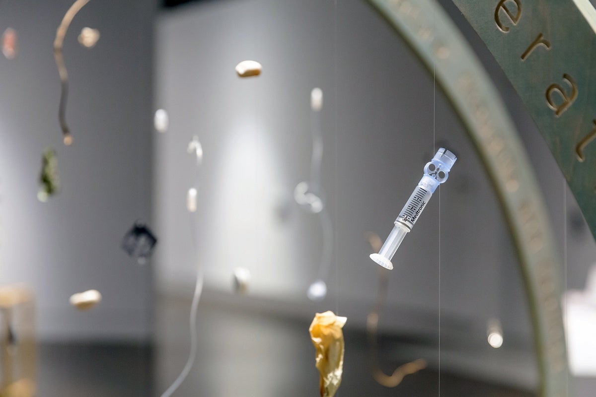 A sculpture of two intersecting circular structures, with words cut into the sides and items hanging in the center of the sculpture. Detail of one of the items shows a syringe.