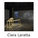 Darkened art gallery with a projection on the back wall, fabric banner with a leaf print hung from twigs and two small tables with organic material, text reads "Clara Laratta".