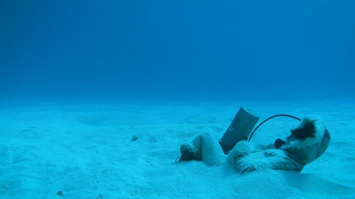 A woman wearing a hooded parka with an oxygen tank and breathing apparatus lays partially buried in sand under the water.
