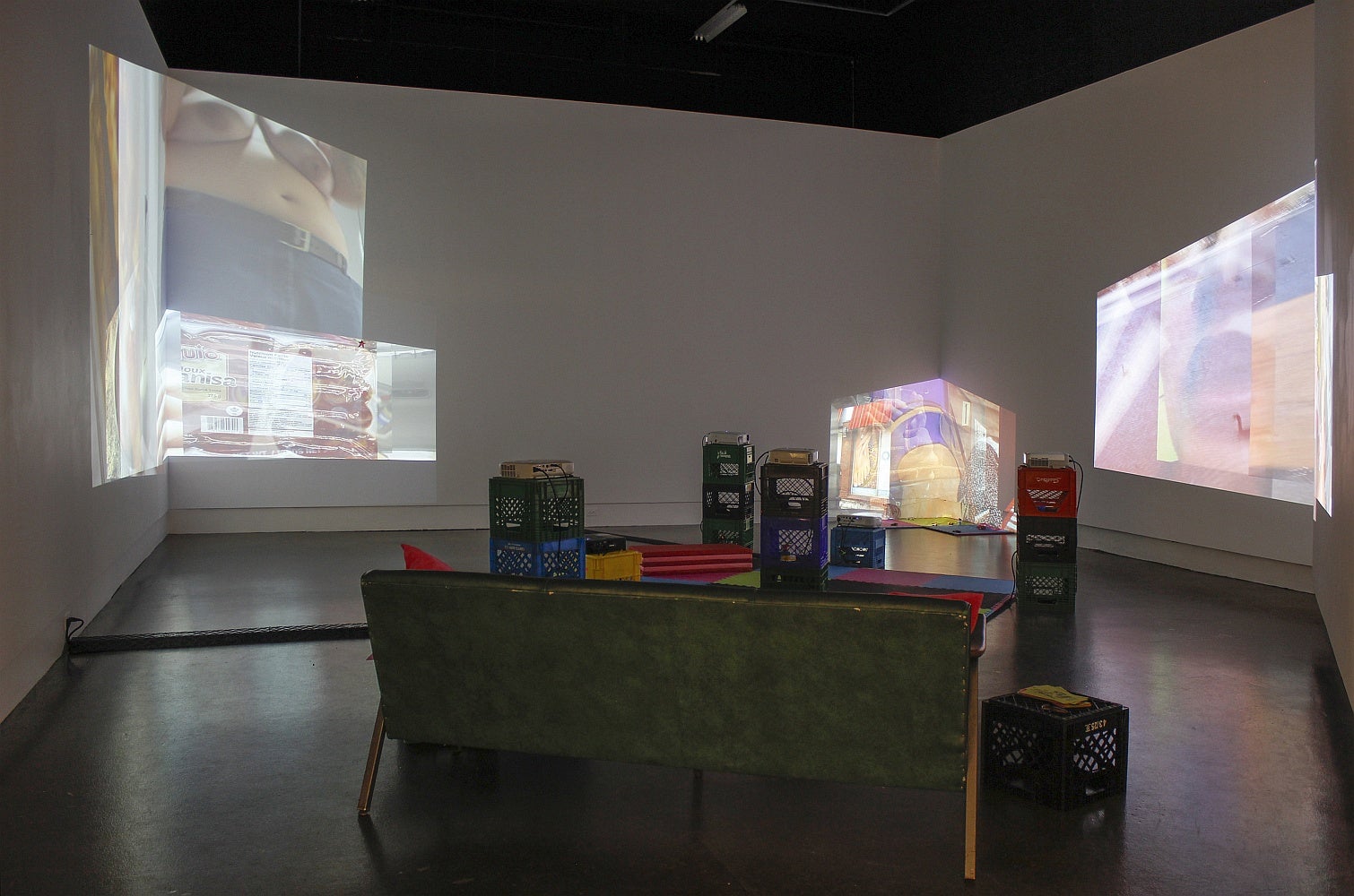 A gallery space with a couch, vinyl mats and multiple projectors set on stacked plastic crates has overlapping projections of videos on several walls.