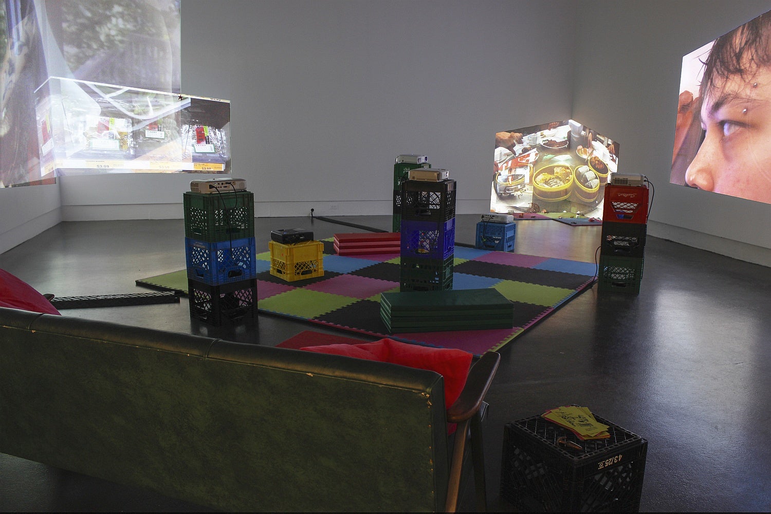 A gallery space with a couch, vinyl mats and multiple projectors set on stacked plastic crates has overlapping projections of videos on several walls.