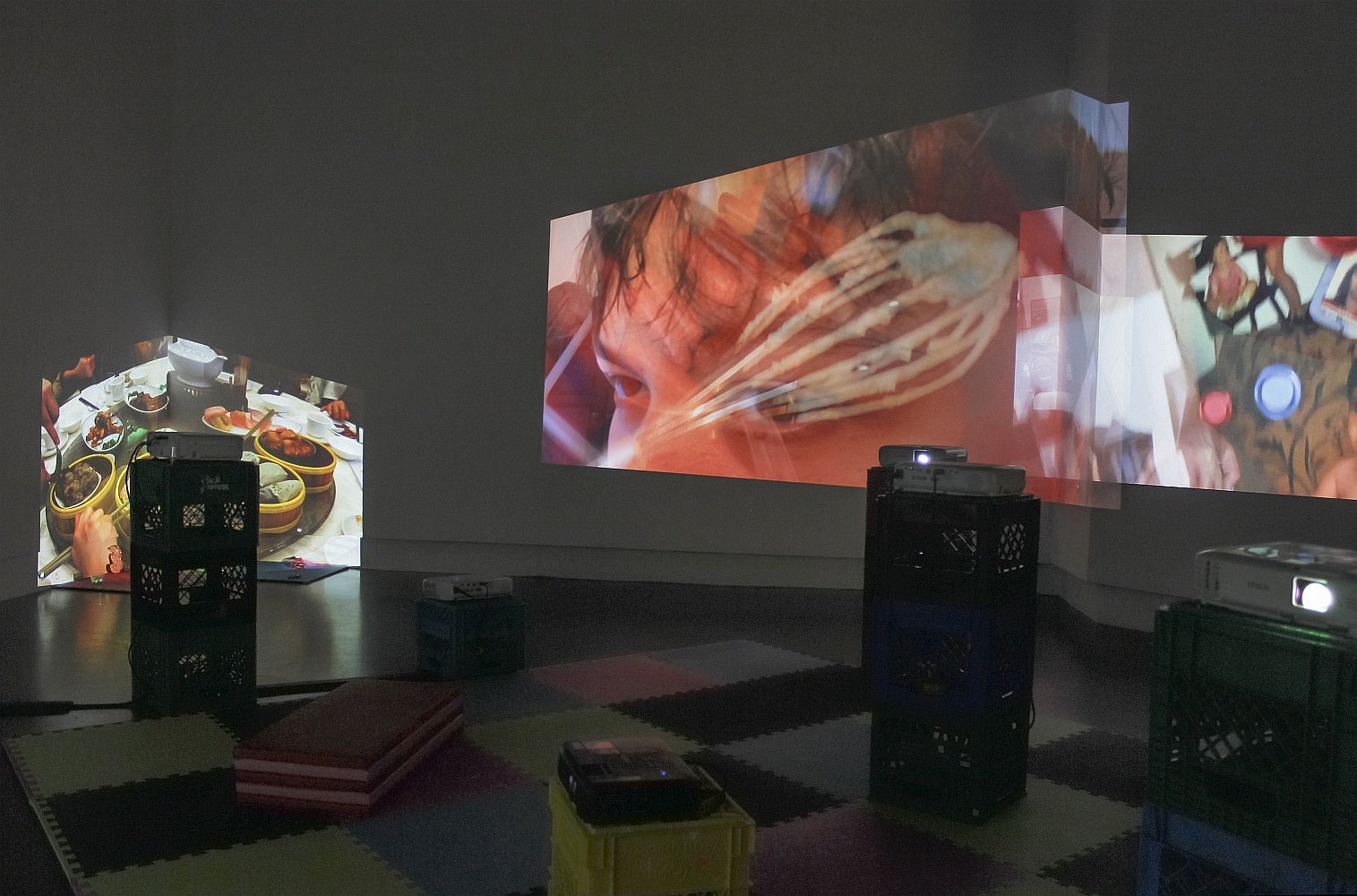 A gallery space with foam floor tiles, vinyl mats and multiple projectors set on stacked plastic crates has overlapping projections of videos on several walls.