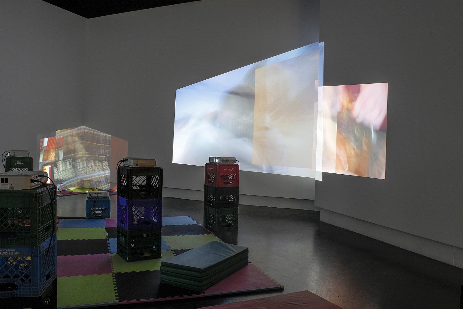 A gallery space with foam floor tiles, vinyl mats and multiple projectors set on stacked plastic crates has overlapping projections of videos on several walls.