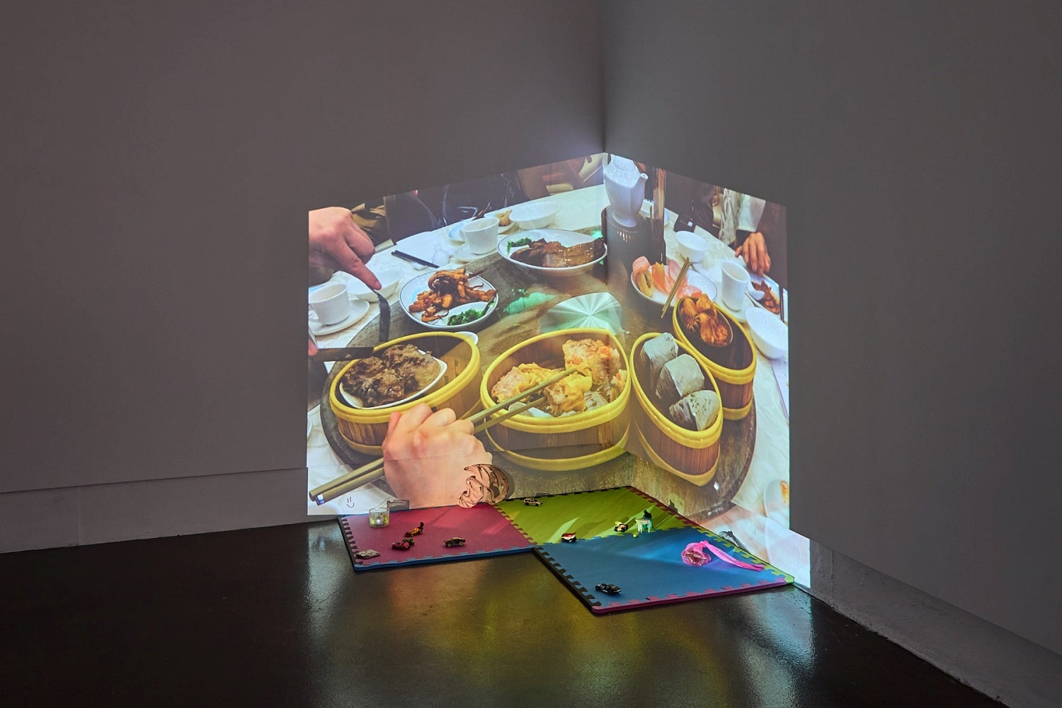 A video is projected into the corner of a room at floor level with toy cars on multi-coloured foam tiles and on the floor