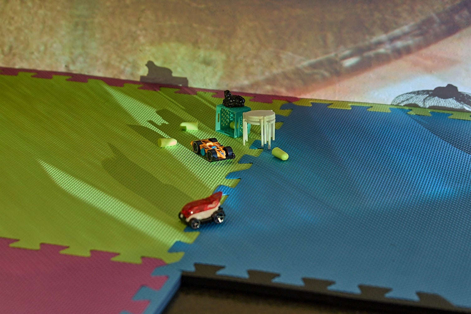 Toy cars and furniture on multi-coloured foam tiles and on the floor with a detail of a video projection on the wall behind.