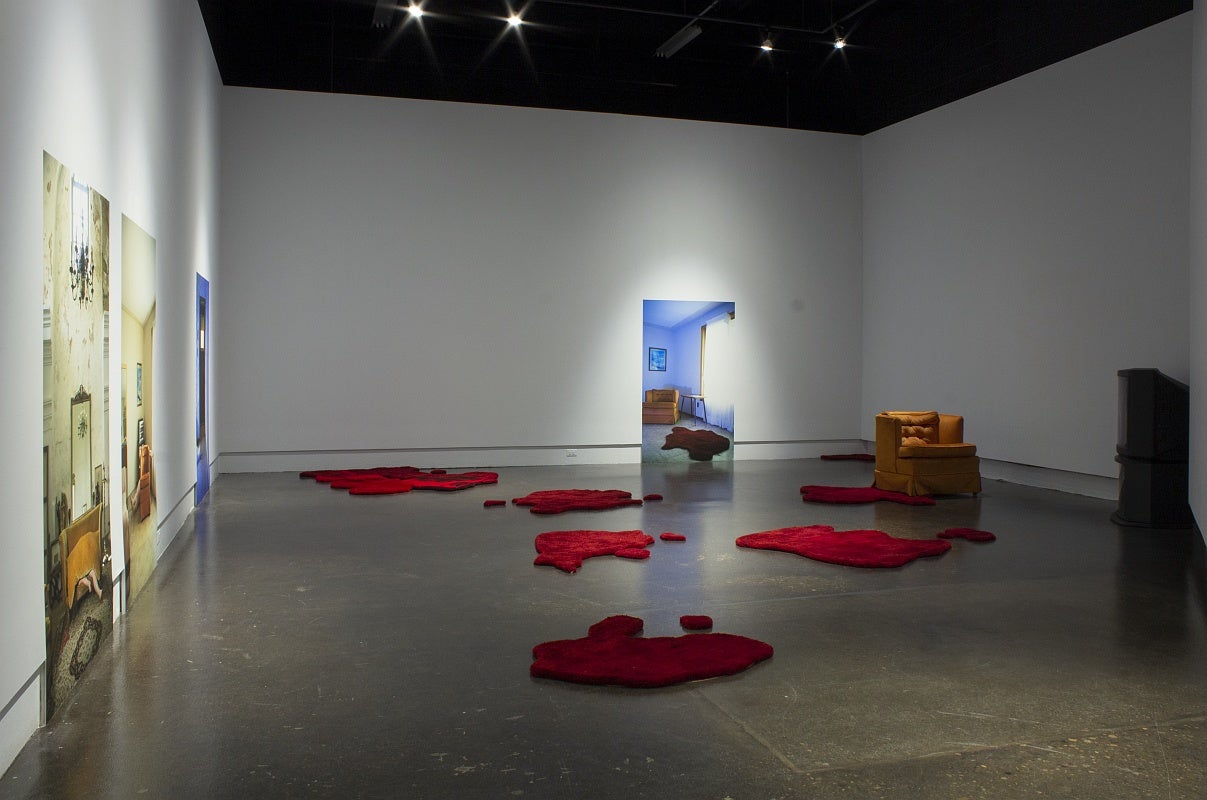 Art installation in a gallery with four large photographs of room interiors, an orange armchair and a vintage television and several irregularly shaped red floor rugs that resemble pools of blood.