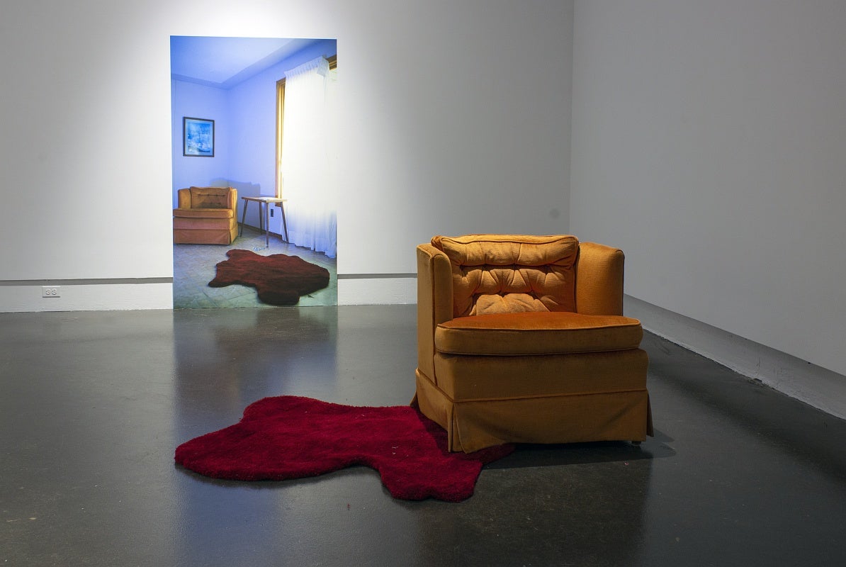 Art installation in a gallery with a large photograph of a living room interior with an orange armchair and red rug. In front is the actual orange armchair and an irregularly shaped red floor rug that resembles a pool of blood.