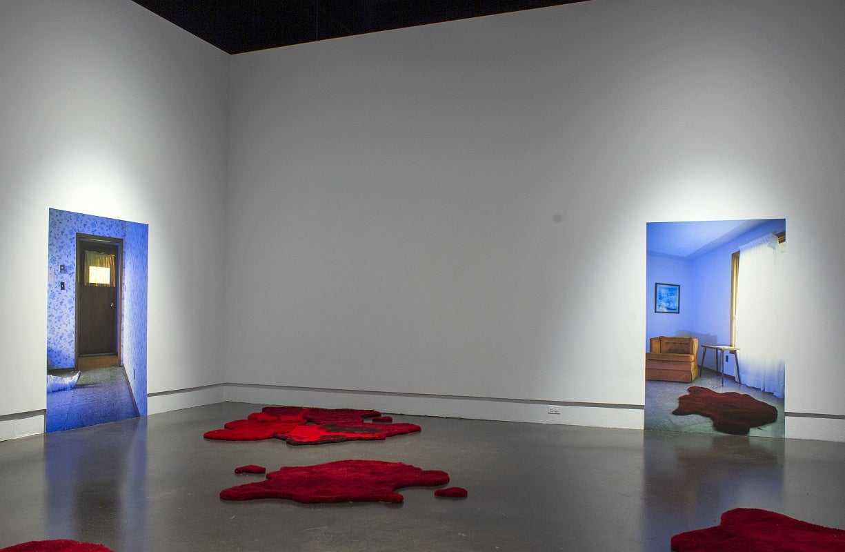 Art installation in a gallery with two large photographs of room interiors, one showing a woman's legs on the floor and other an orange armchair and red rug. On the gallery floor are several irregularly shaped red floor rugs that resemble pools of blood.