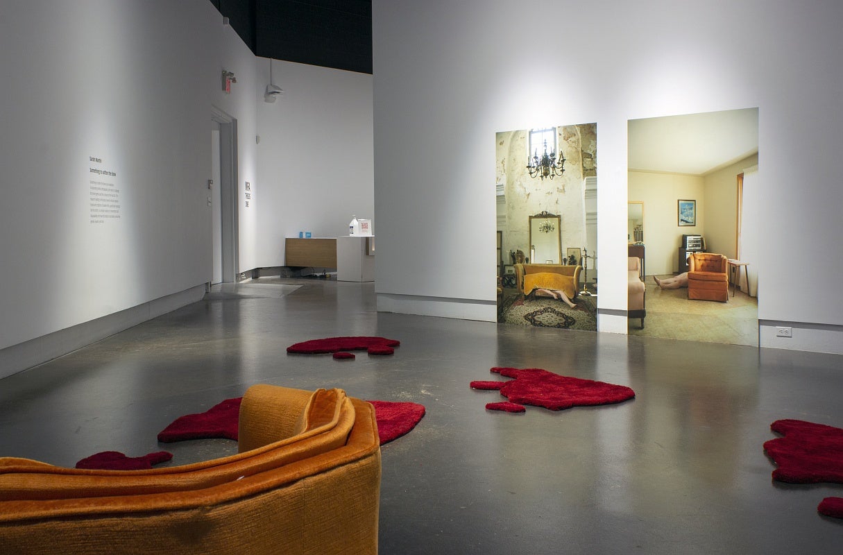 Art installation in a gallery with two large photographs of room interiors, one showing a white woman’s body under a sofa cushions and in the second she is lying on the linoleum floor between an old orange armchair and a stereo cabinet. On the gallery floor are several irregularly shaped red floor rugs that resemble pools of blood and in the front corner is the orange armchair.