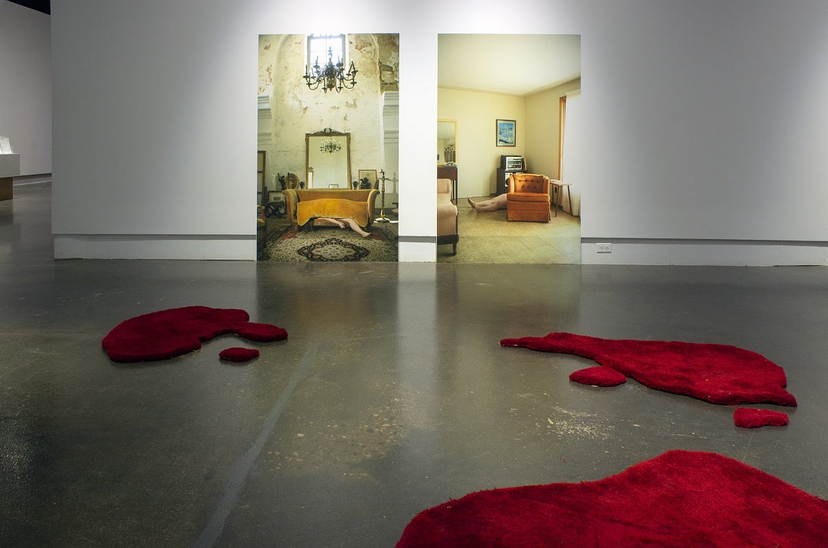 Art installation in a gallery with two large photographs of room interiors, one showing a white woman’s body under a sofa cushions and in the second she is lying on the linoleum floor between an old orange armchair and a stereo cabinet.