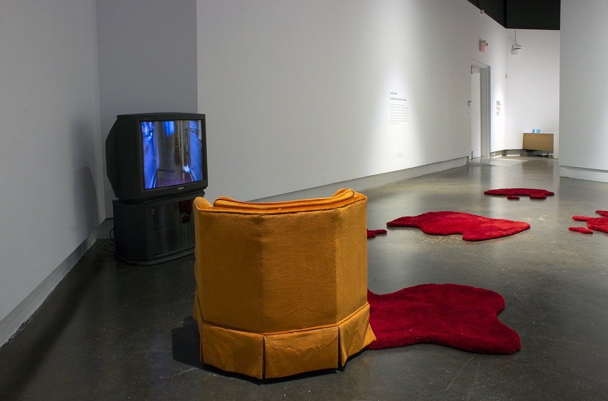 An art gallery with several irregularly shaped red floor rugs that resemble pools of blood and in the left corner is the orange armchair and a 1980's television set displaying a hallway.